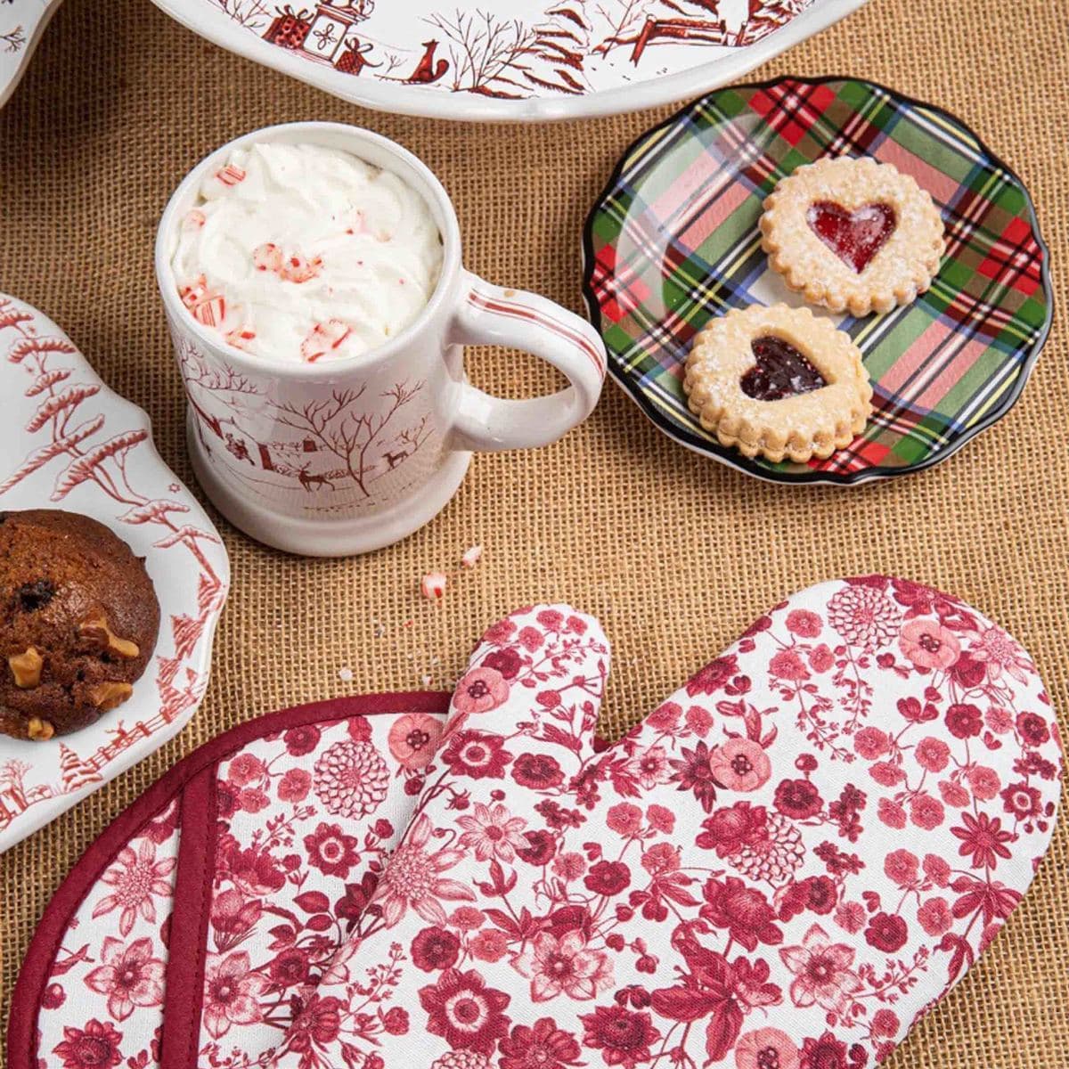 Mug with whipped cream, cookies on a plaid plate, and floral oven mitt on a textured surface.