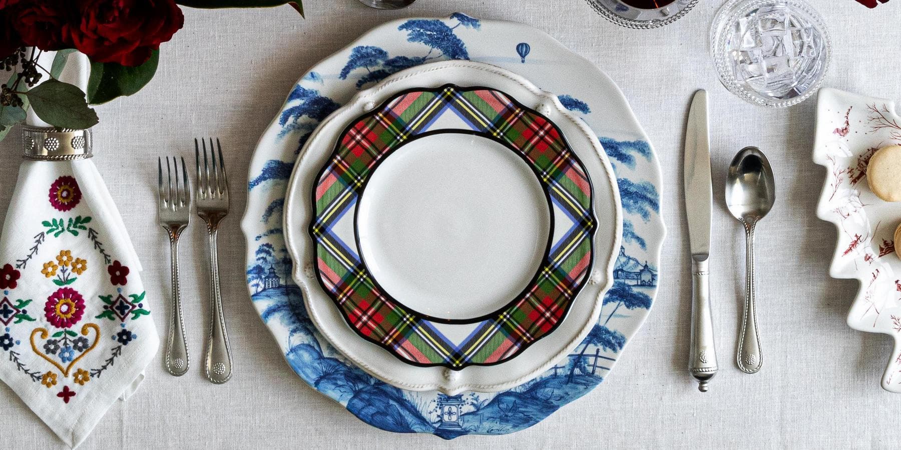 Dining table setting with a plaid plate, silverware, and decorative items on a white tablecloth.