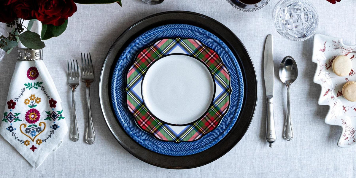 A styled table setting featuring a decorative place setting with a white plate, a blue braided charger, a red and green tartan patterned plate, silver cutlery, a white embroidered napkin, and a white floral dessert plate with cookies.