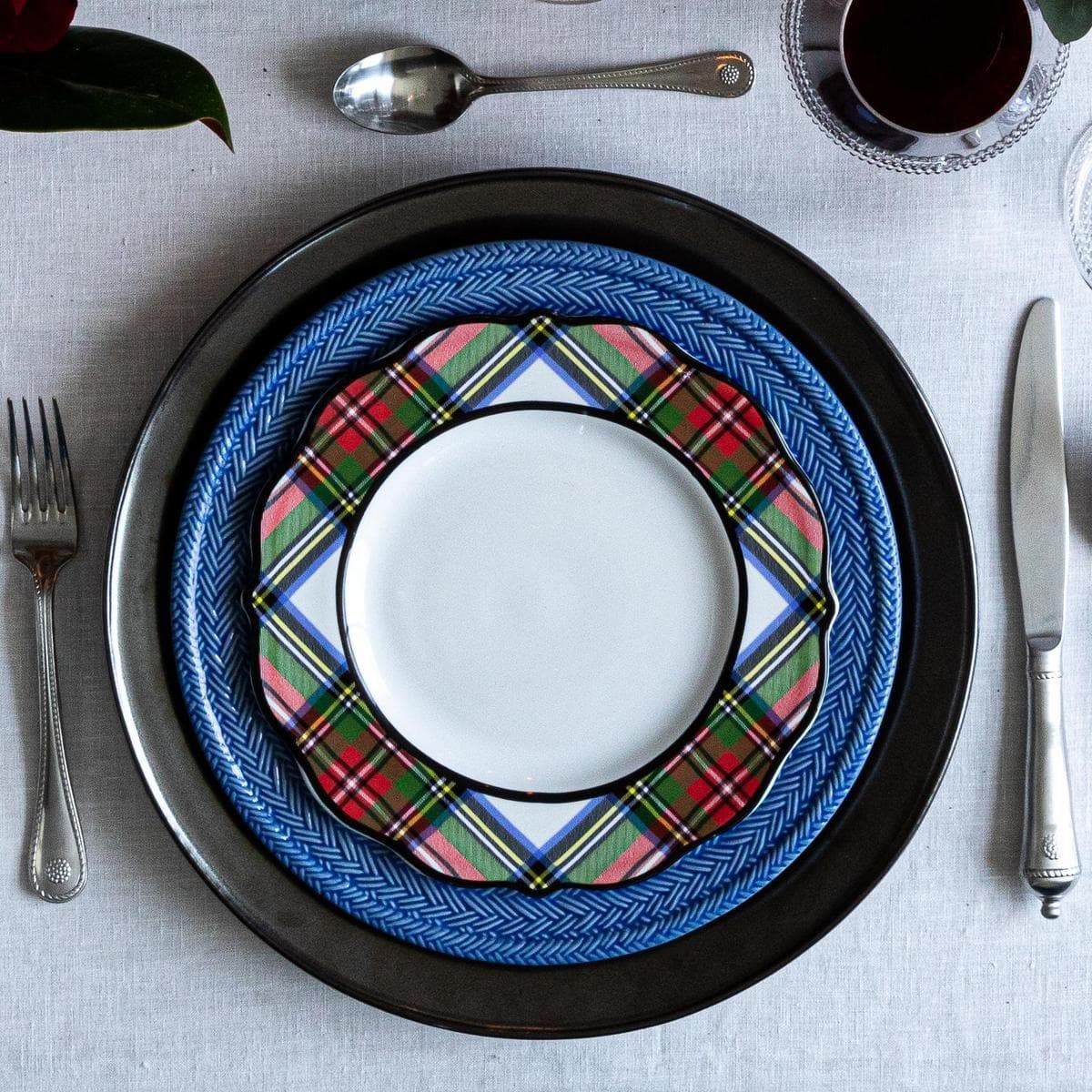 A styled table setting featuring a stack of three decorative ceramic plates: a black charger plate, a blue and tartan-patterned plate, and a white center plate, surrounded by silver cutlery and a glass of red wine on a light gray tablecloth.