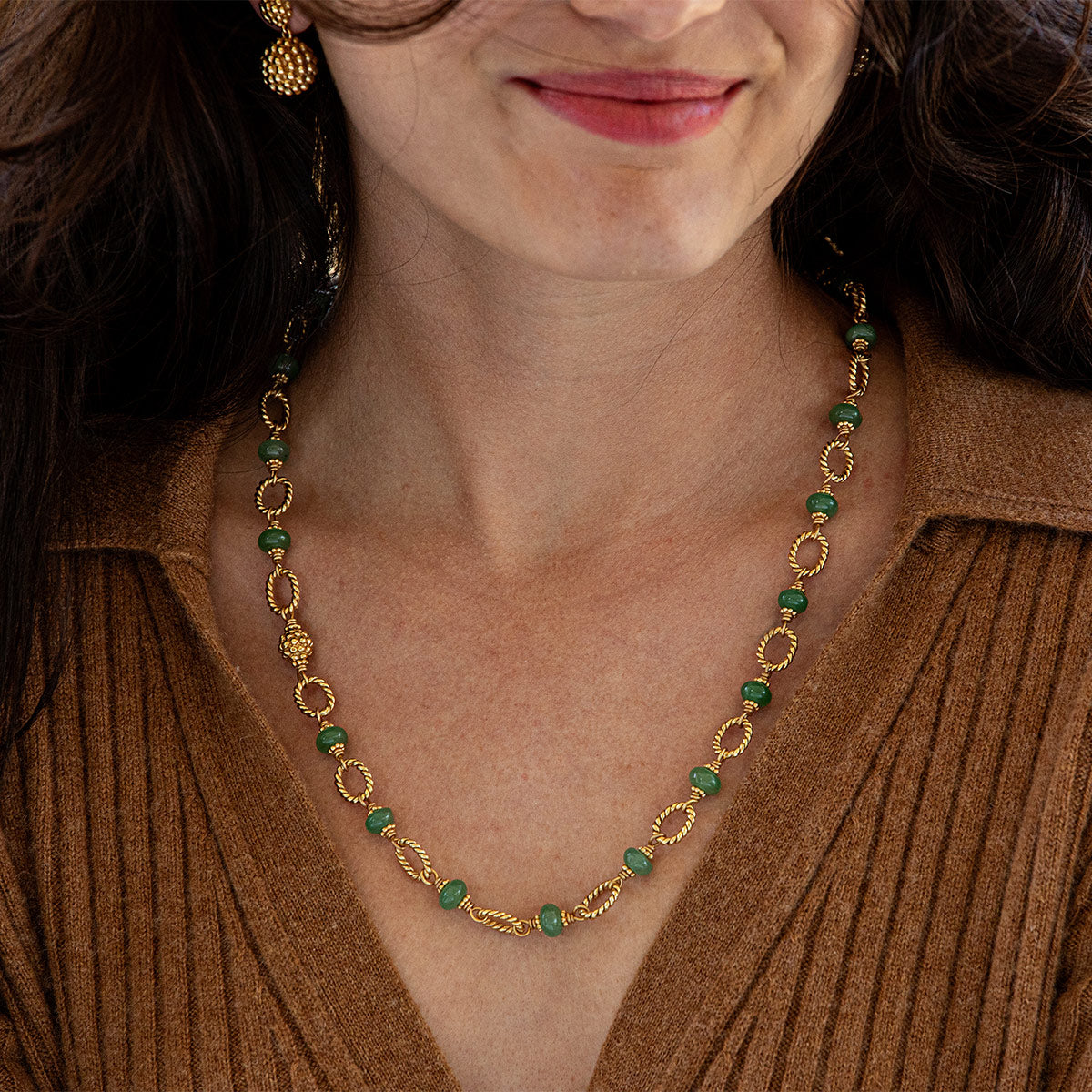 A close-up of a woman wearing a gold necklace with green stone accents and matching dangling earrings, paired with a brown ribbed knit top.