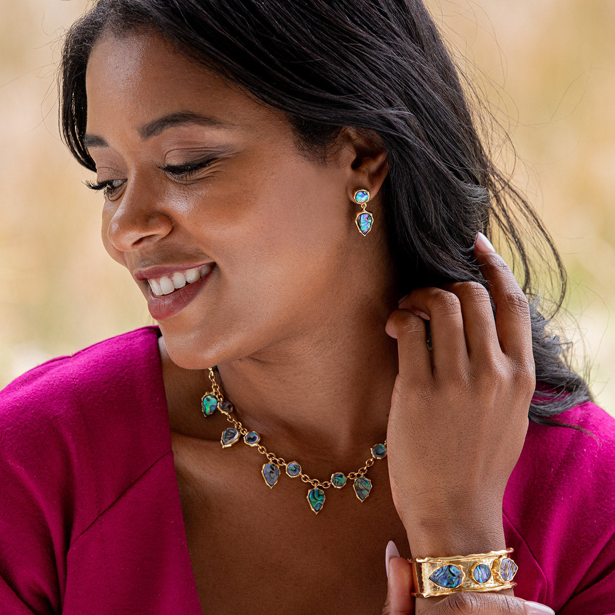 A woman wearing a gold jewelry set with turquoise-blue stones, including a necklace, matching drop earrings, and a cuff bracelet, paired with a bright magenta top against a soft blurred background.