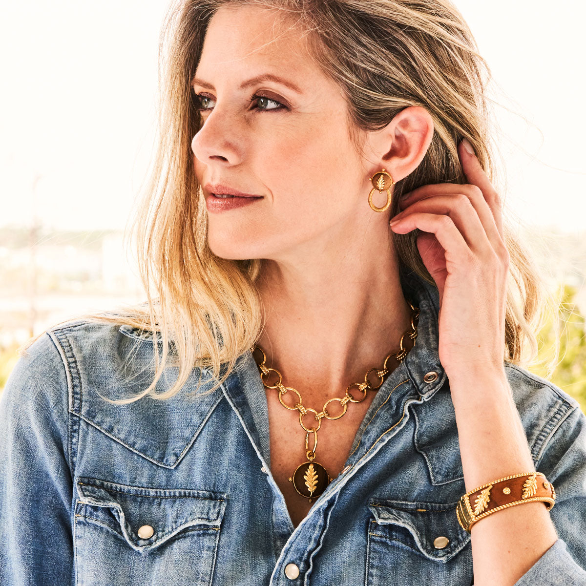 Woman wearing gold jewelry including earrings, necklace, and bracelet with a blurred outdoor background