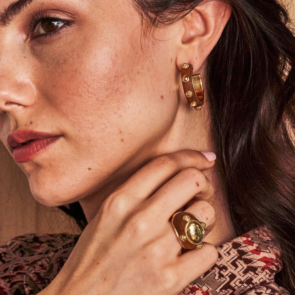 Close-up of a woman wearing gold hoop earrings and a ring, with a blurred background.