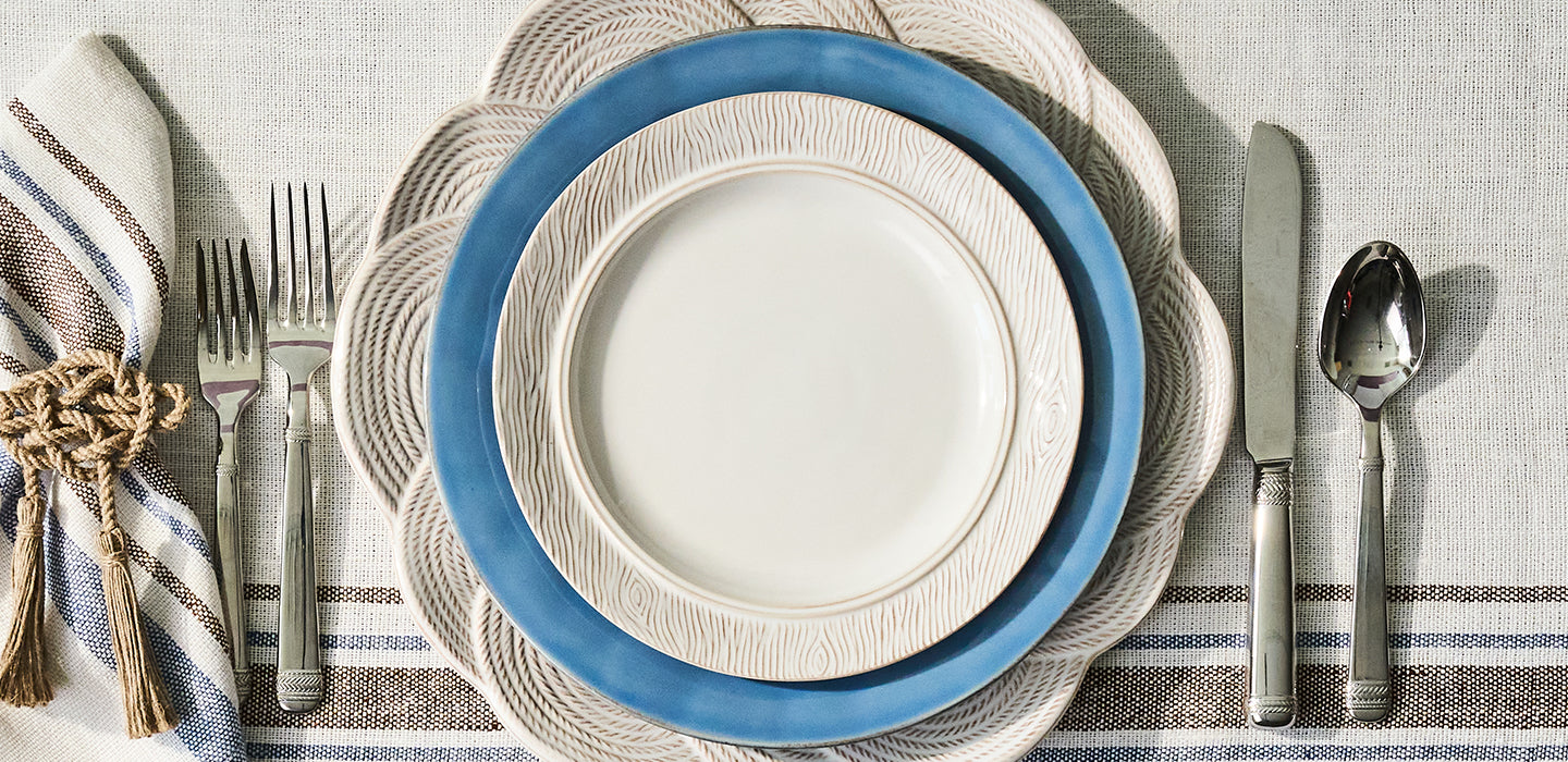 Dining table setting with blue and beige plates, silverware, and striped napkins on a textured tablecloth.