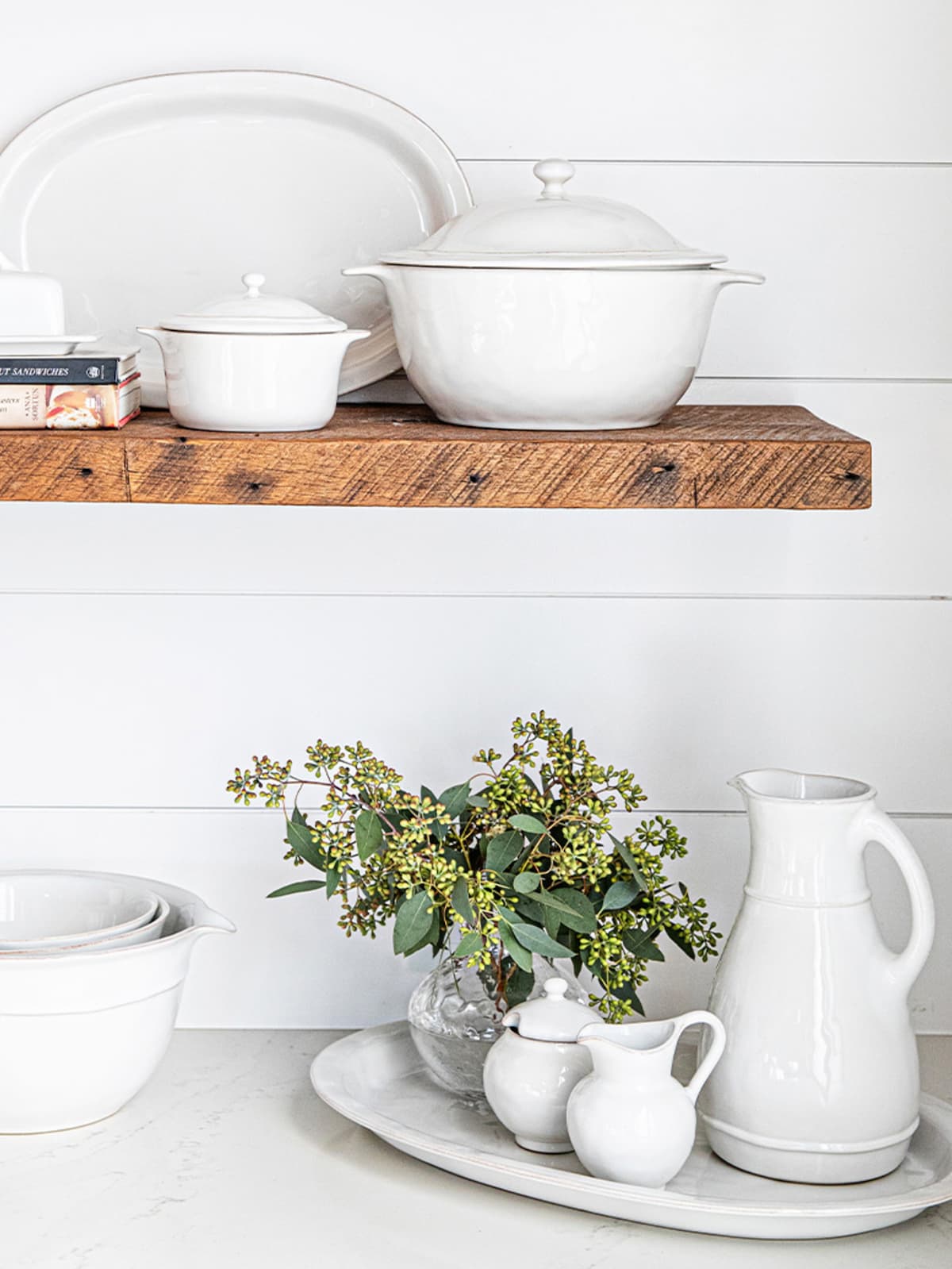 White ceramic dishes on a wooden shelf with a vase of greenery on a white surface.