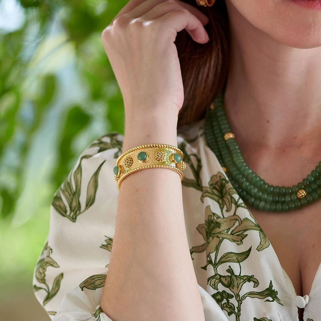Close-up of a person wearing gold bracelet and green beaded necklace with a blurred natural background